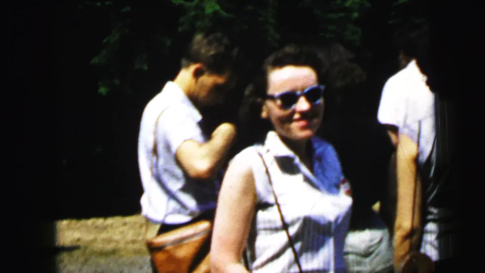 Woman in blue cat-eye sunglasses smiling beside a man in summer clothing, third figure at right, trees in background; 8mm home movie, c. 1964.