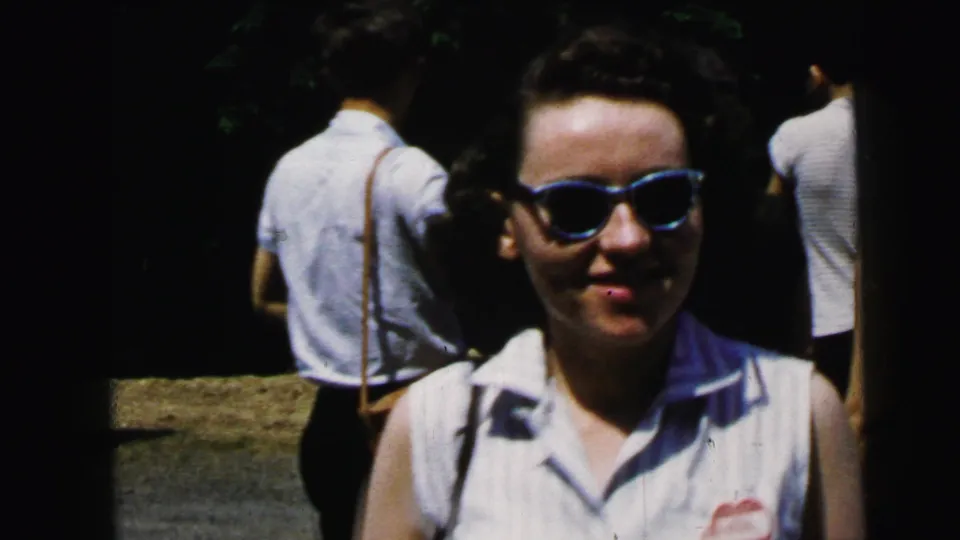 Woman in blue cat-eye sunglasses and sleeveless collared blouse smiling directly at camera; background figures suggest a public outdoor venue, 8mm, c. 1964.