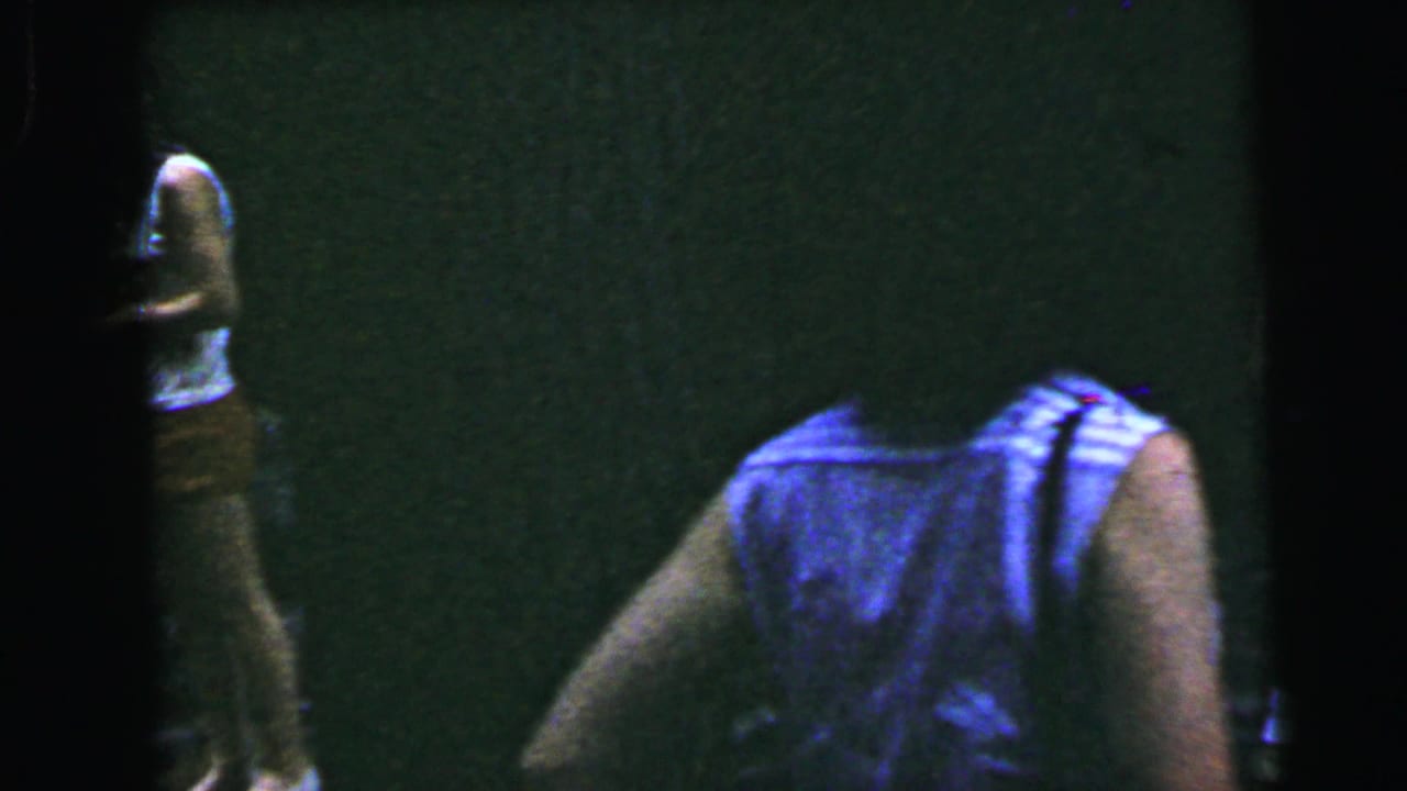 Opening frame of a 1964 8mm home movie — a partial figure barely visible at left edge against a near-black interior; severe underexposure limits detail.
