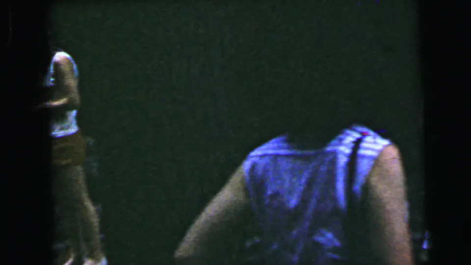 Opening frame of a 1964 8mm home movie — a partial figure barely visible at left edge against a near-black interior; severe underexposure limits detail.