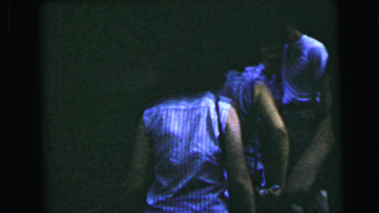 A person seen from behind in a blue sleeveless top, set against a very dark interior on 8mm film.