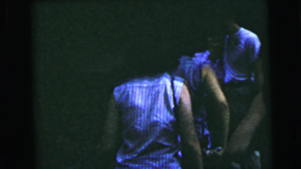 Rear view of a woman in a blue sleeveless top shot on 8mm home movie film in a dark interior, consistent with the 1964 mid-summer domestic setting.