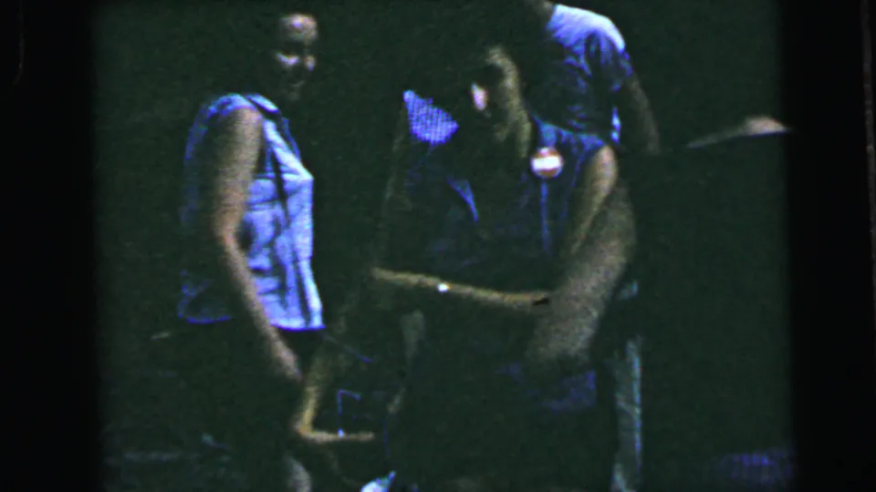 Two women in close confrontation in an unlit indoor space on 8mm home movie film circa 1964; gestural body language and proximity indicate an active dispute.