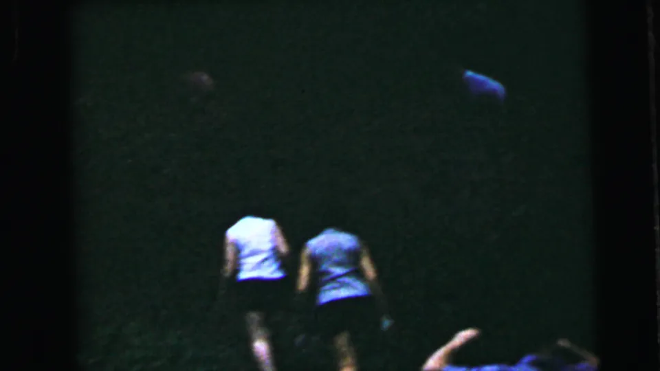 Wider shot of two women from behind in a dark interior on 8mm home movie film circa 1964; both in sleeveless tops, framing suggests movement or separation.