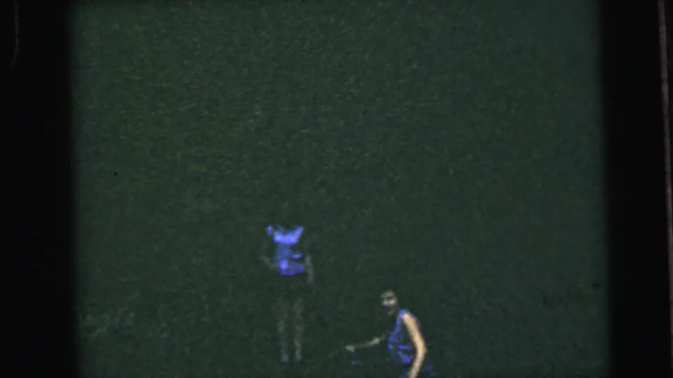 Two women in a darkly lit wooded setting, one appearing to bend or crouch, in a mid-1960s 8mm home movie clip.