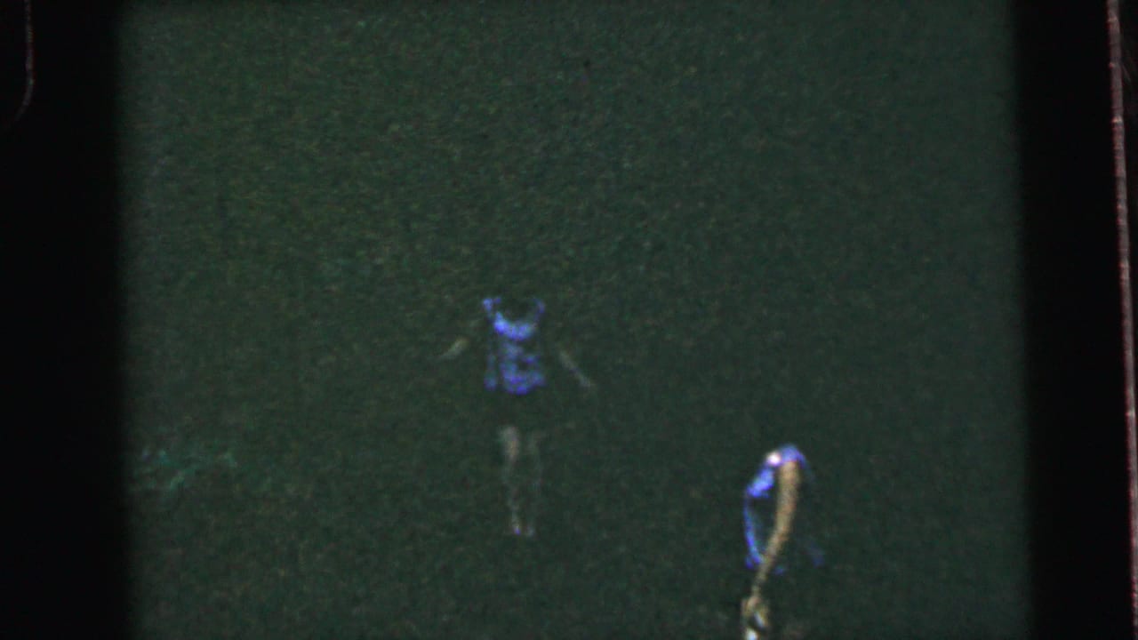 Two female runners in blue sleeveless tops and dark shorts race across an open field, possibly Camden, New Jersey, circa 1964; heavy grain and greenish cast from 8mm film.