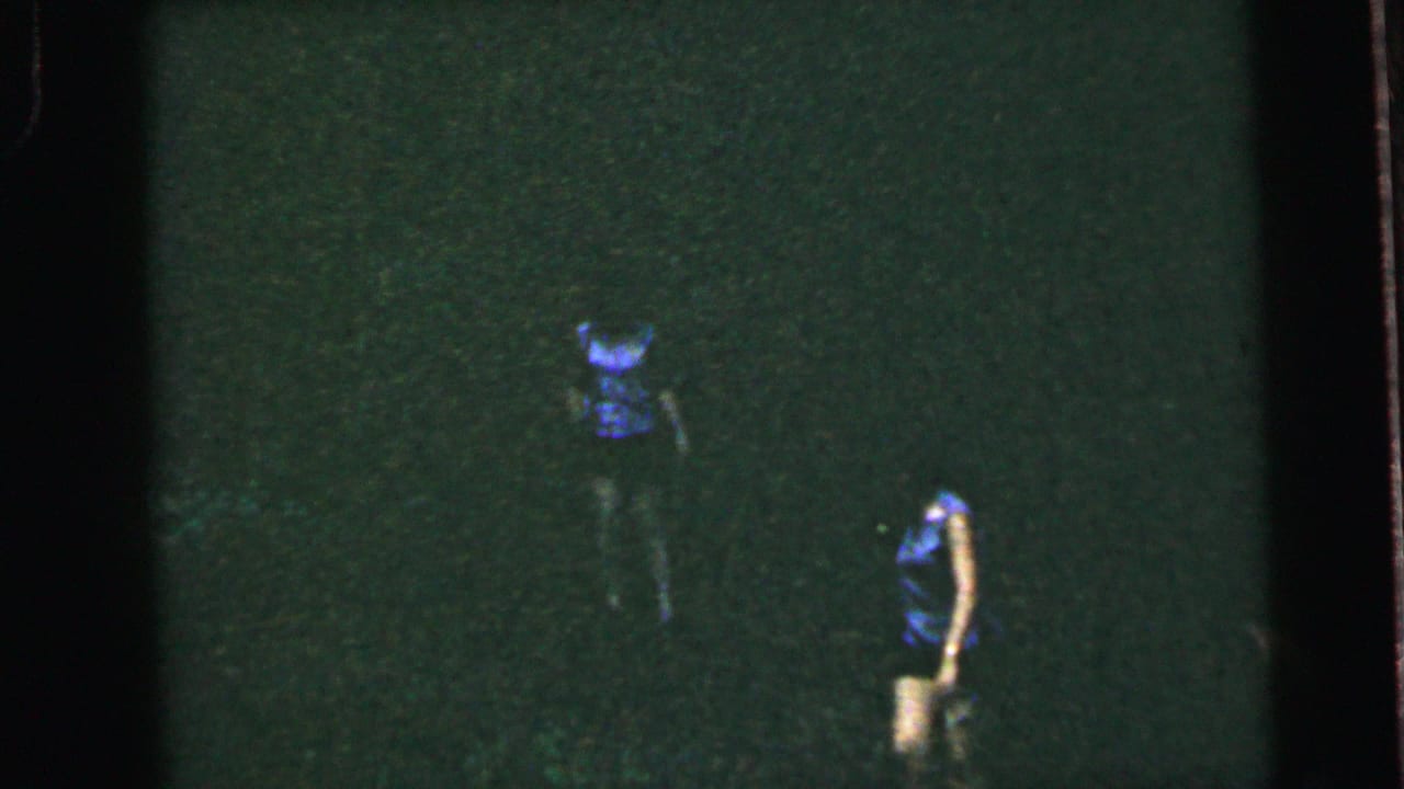 Two female runners on a dark open field — one in blue top, another in a lighter garment to the right — on 8mm film with pronounced grain and green color shift, circa 1964.