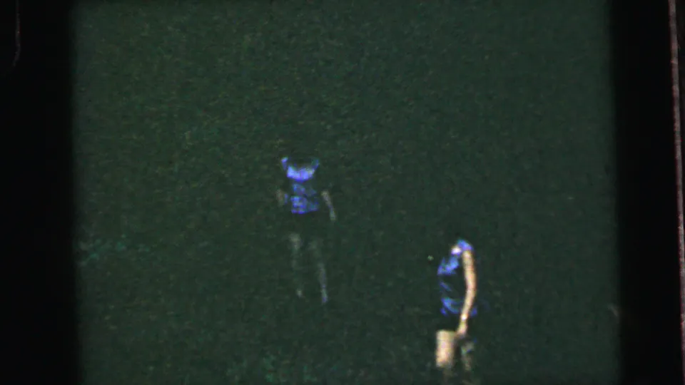 Two female runners on a dark open field — one in blue top, another in a lighter garment to the right — on 8mm film with pronounced grain and green color shift, circa 1964.