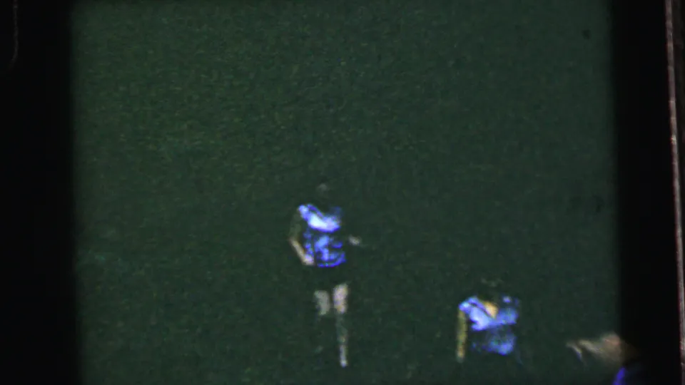 Two figures running across a dark, heavily underexposed grass field on 8mm film with deep green color cast, possibly Camden, New Jersey, circa 1964.