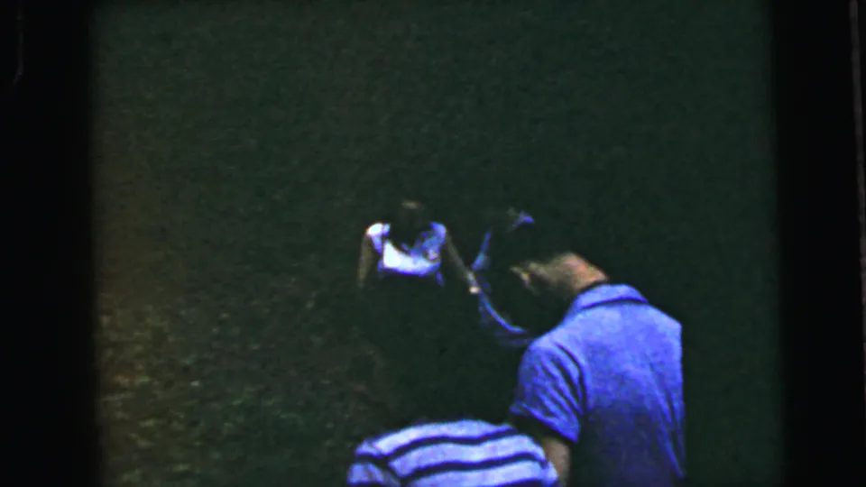 Adult male in a blue-and-white horizontal striped shirt stands in the foreground at the field edge with arm raised, while runners appear in the dark background on underexposed 8mm film, circa 1964.