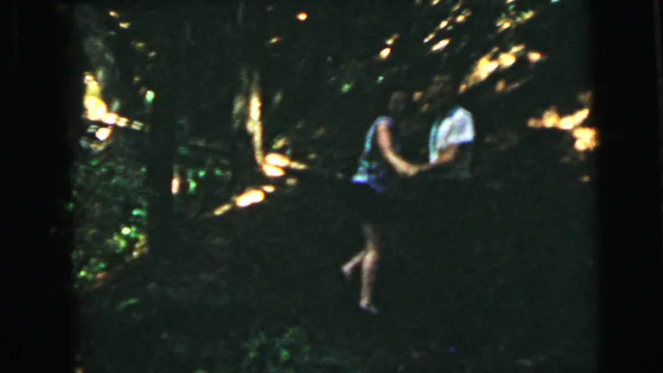 A figure in light-colored clothing moves through dense forest with patches of warm golden sunlight breaking through the tree canopy, 8mm footage circa 1964.