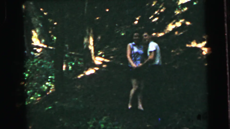 A woman in a patterned sleeveless top and light skirt poses toward the camera in a sun-dappled forest clearing, a second figure partially visible behind her, 8mm home movie circa 1964.