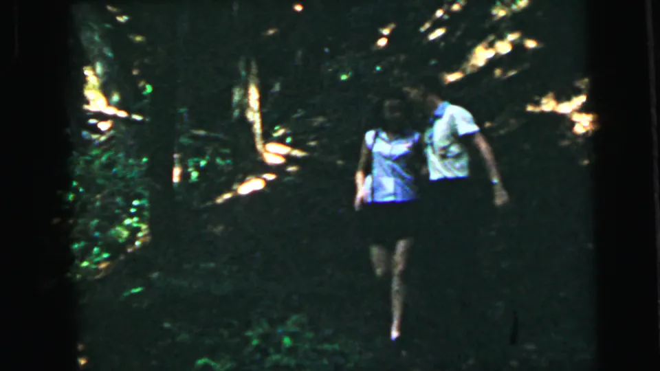Young couple in summer clothing walking through a wooded setting, mid-distance shot on 8mm home movie film, circa 1964.