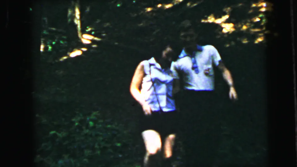 Young woman in sleeveless top and young man in white short-sleeve shirt walking toward camera through summer woodland, 8mm home movie circa 1964.