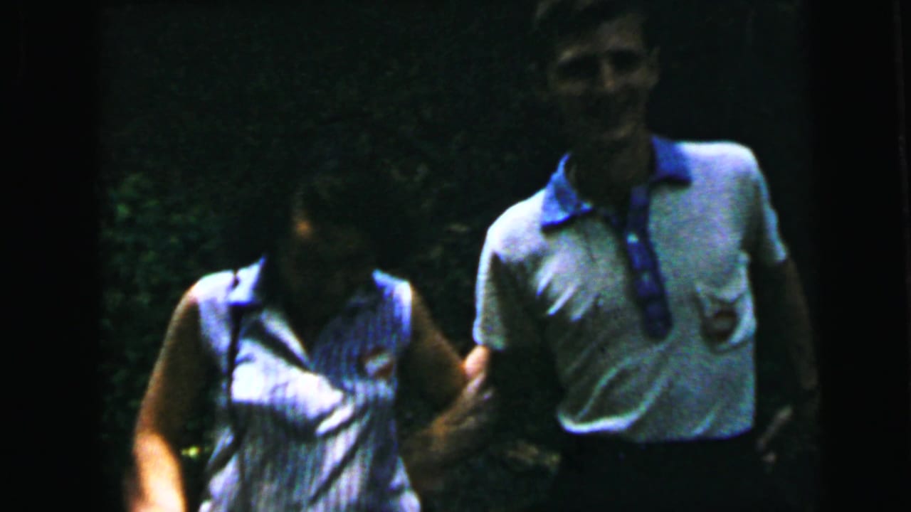 Young man and woman walking together through a wooded area, facing the camera, in casual early 1960s summer clothing.