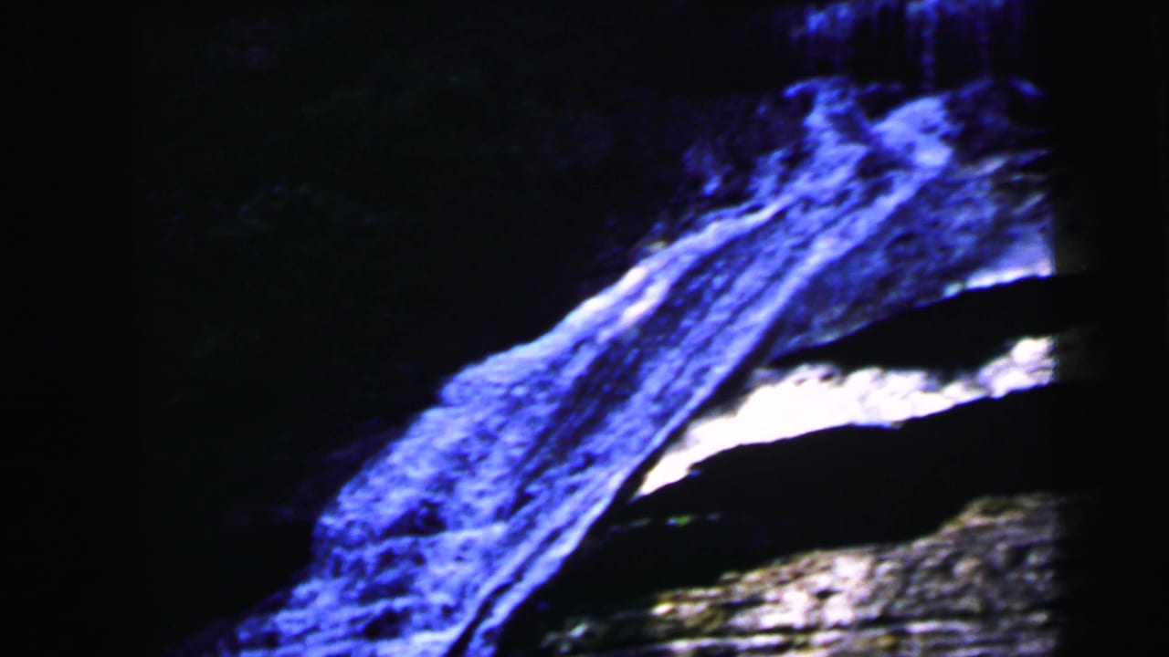 8mm film close-up of water flowing diagonally over dark rock, heavily blue-shifted, from a 1960s natural-attraction outing.