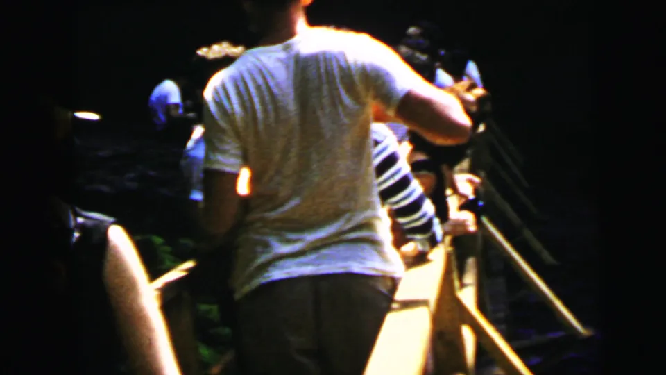 1960s tourists on a wooden observation walkway in a dark gorge; man in white t-shirt and other visitors seen from behind, warm light source at upper left.