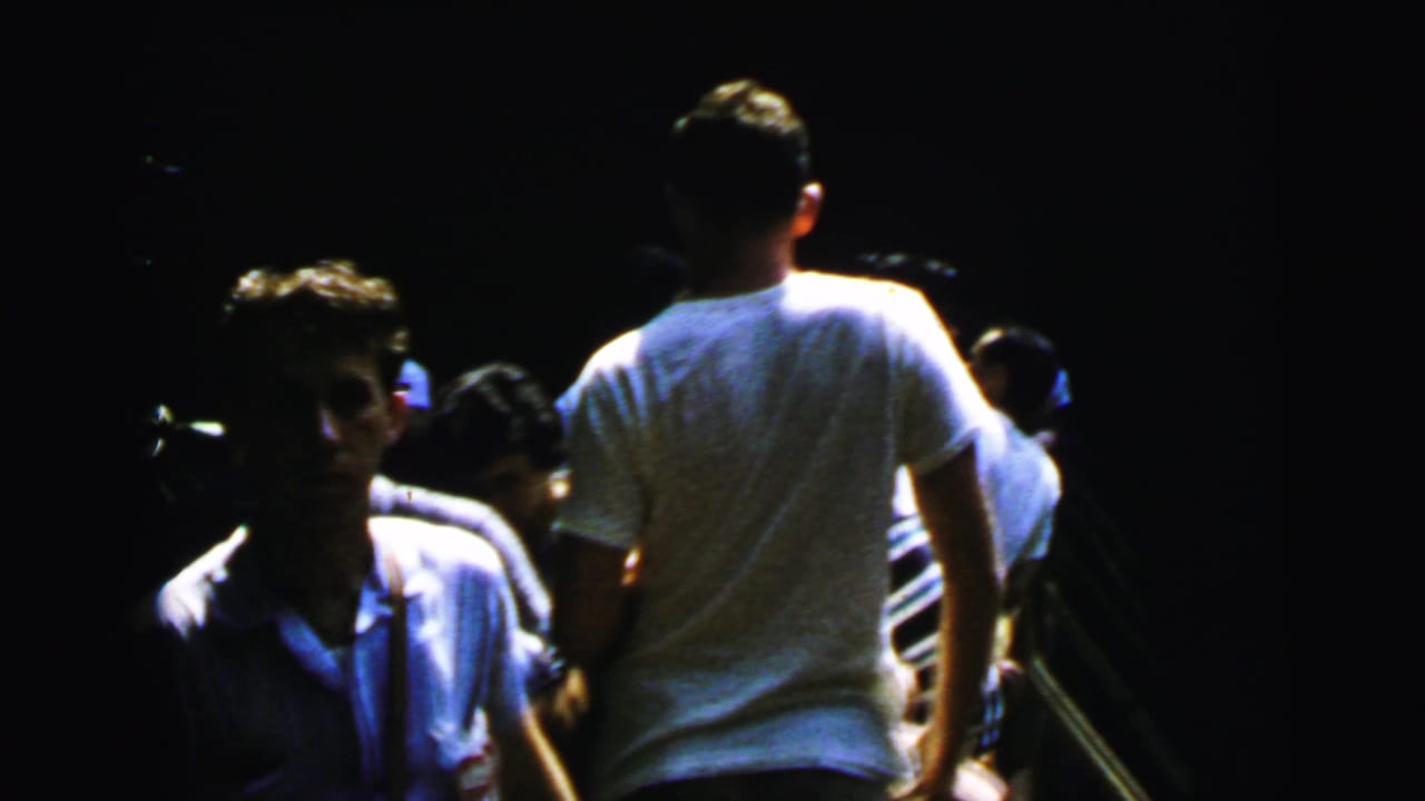 Group of young adults walking through a dark shaded outdoor setting; a man in white t-shirt seen from behind leads the group.