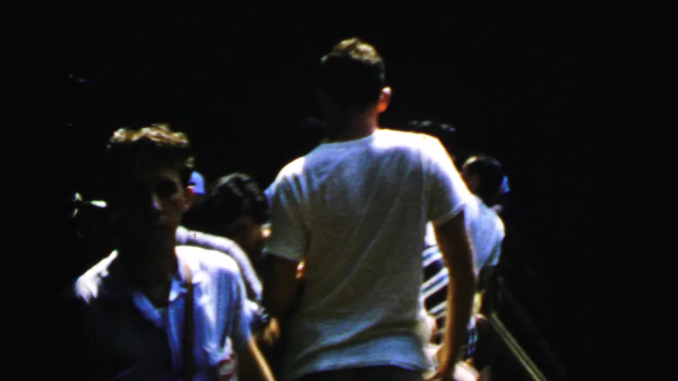 1964 8mm home-movie frame: group of young adults walking through a dark outdoor setting, man in white t-shirt prominent from rear, others in casual summer clothing.