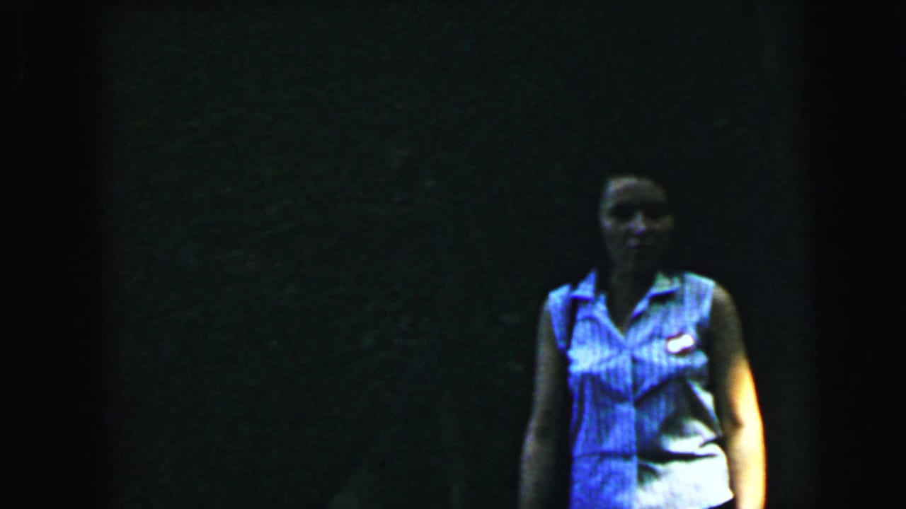 Young woman in a blue-and-white striped sleeveless uniform with a small chest badge, standing in a dark outdoor setting; 1964 8mm home-movie footage, Camden, New Jersey area.