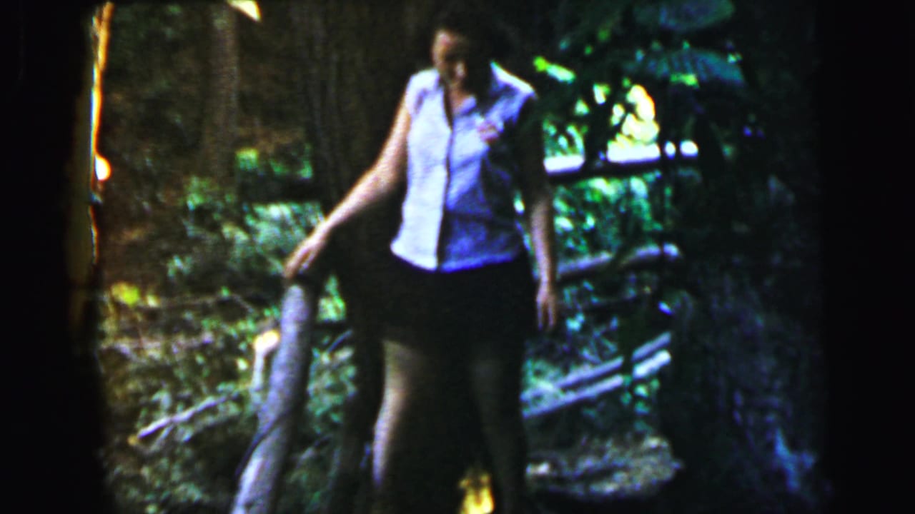 Woman in light sleeveless blouse and dark skirt beside a large tree, wooden bridge or railing structure in background, wooded setting, circa 1964.