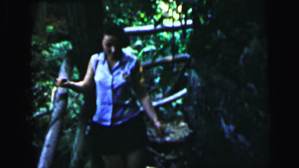 Woman in light sleeveless blouse and dark skirt navigating a log or plank structure amid dense woodland, 8mm home movie, circa 1964.