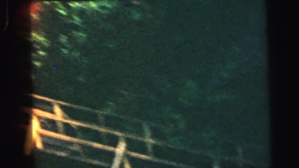 Underexposed 8mm home movie frame showing bridge railings in near-darkness with a red-orange light flare at the left edge; no figures discernible.