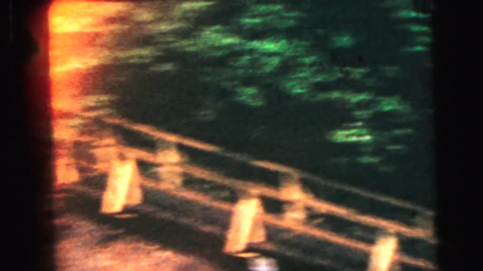 8mm home movie from 1964 showing bridge planks and cross-rail structure lit by warm orange-amber light from below, viewed from a low angle with dense dark foliage above.