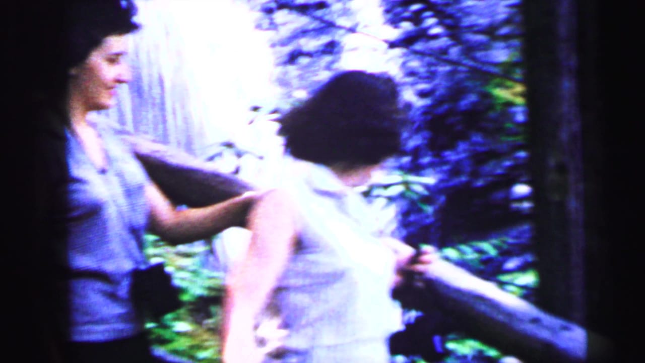 Two women near a waterfall, one in a white sleeveless top gripping a wooden rail, the other with dark hair beside her