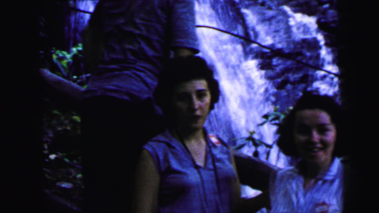 Two women stand near a cascading waterfall on a rocky wooded face; one smiles broadly toward camera, the other stands more contemplatively in a sleeveless top.