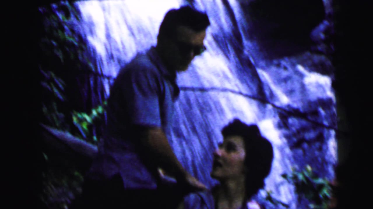 A man in a short-sleeve shirt stands behind a dark-haired woman near a cascading waterfall; she looks upward or to the side.