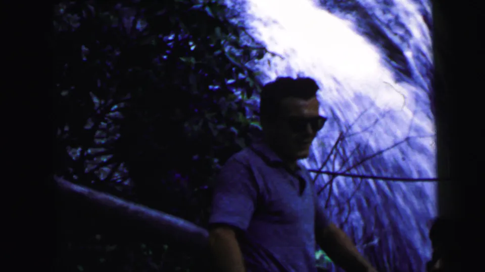 Man in sunglasses and short-sleeve shirt at a wooded waterfall, circa 1964 8mm home movie; heavy corner vignetting and blue-purple color shift visible.