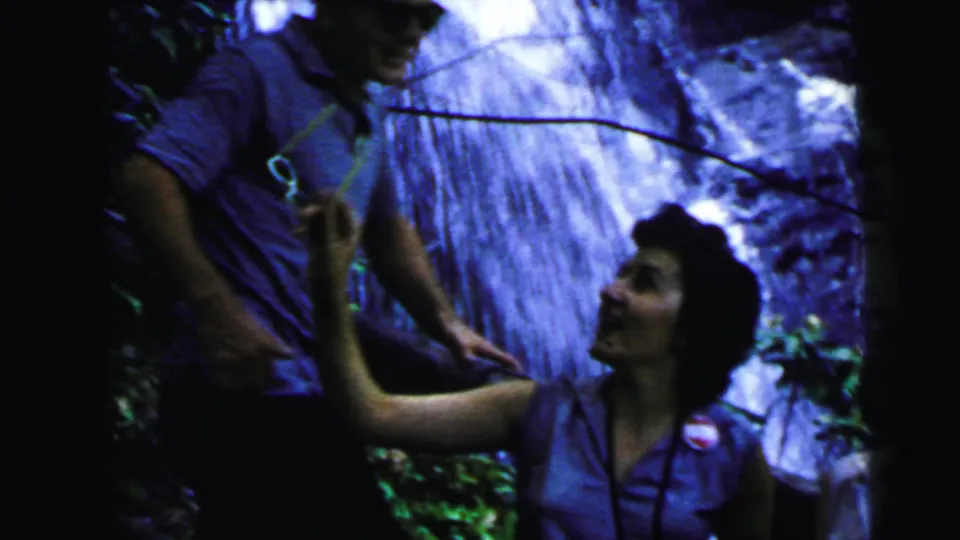 Man and woman interact animatedly before a cascading waterfall, circa 1964 8mm vacation footage; man gestures upward, colorful item near his shoulder, foliage surrounds them.