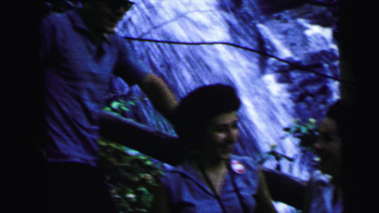 Man's torso and raised arm with a woman near a rocky waterfall; blue-purple toned 8mm film.