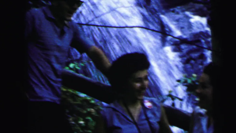 A man's torso and arm partially visible alongside a woman at a rocky waterfall, 8mm home movie with blue-purple color shift, attributed New Jersey, 1964.