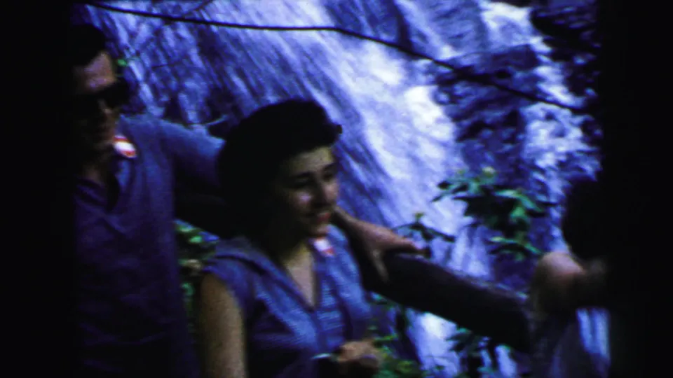 Man in sunglasses gesturing beside a smiling woman at a cascading waterfall, third figure at right frame edge; 8mm home movie attributed New Jersey, 1964.