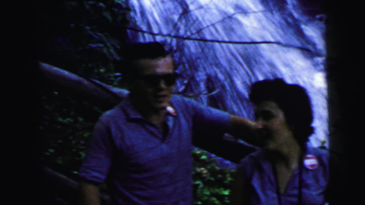 A man in early-1960s casual dress and dark sunglasses stands beside a dark-haired woman at a forest waterfall, captured on 8mm home movie film circa 1964.