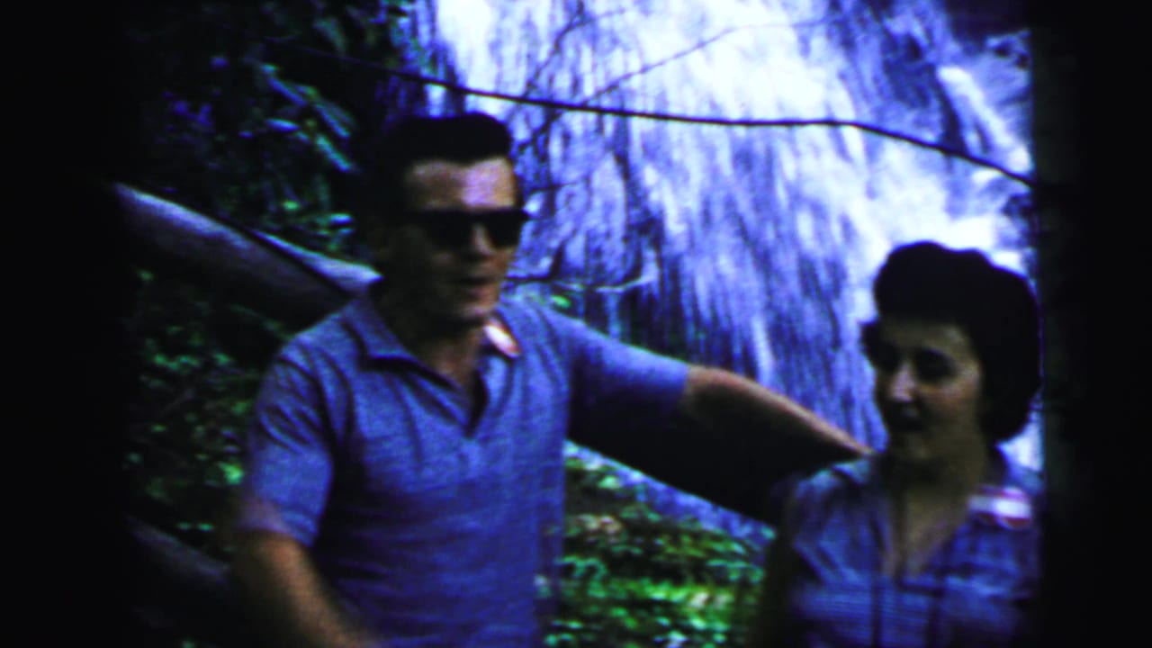 A man gestures toward a large cascading waterfall as a woman looks on, shot on 8mm home movie film consistent with early-1960s footage, possibly New Jersey, 1964.