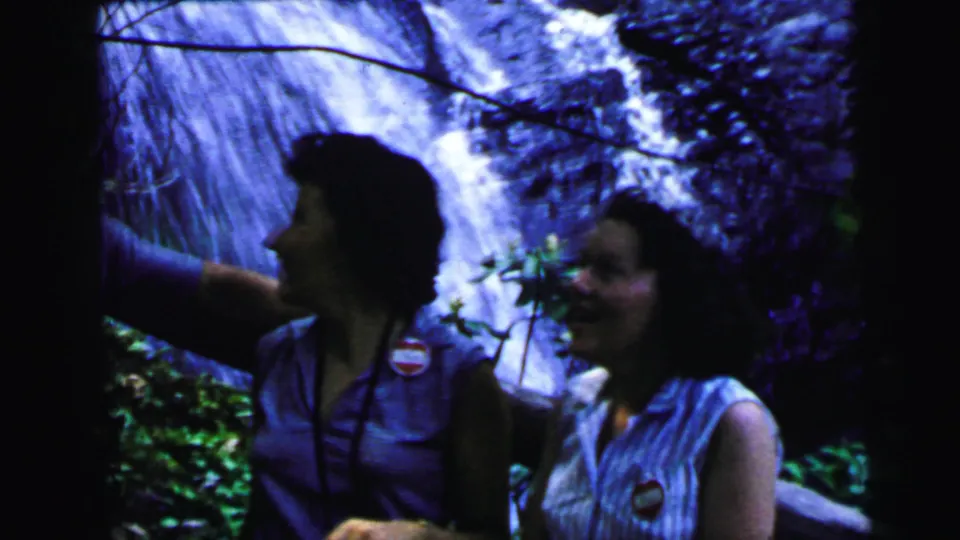 A dark-haired woman in a sleeveless top wearing a circular pin badge stands close to camera at a wooded waterfall, 8mm home movie circa 1964.