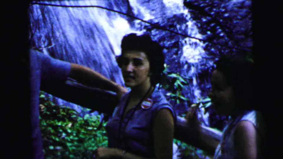 A woman in a sleeveless top with a circular badge stands at a forest waterfall with other family members partially in frame, 8mm home movie circa 1964.