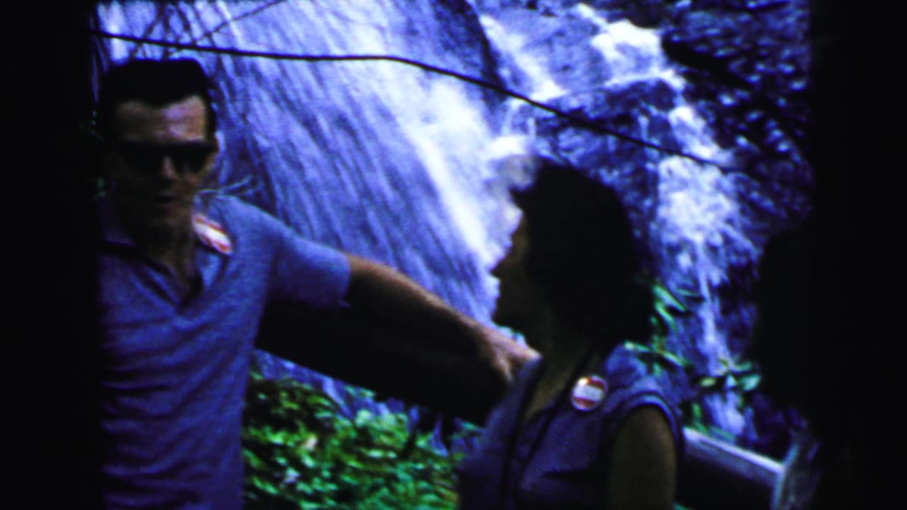 Man in sunglasses and blue polo shirt gesturing beside a woman in a sleeveless top, large cascading waterfall in background.