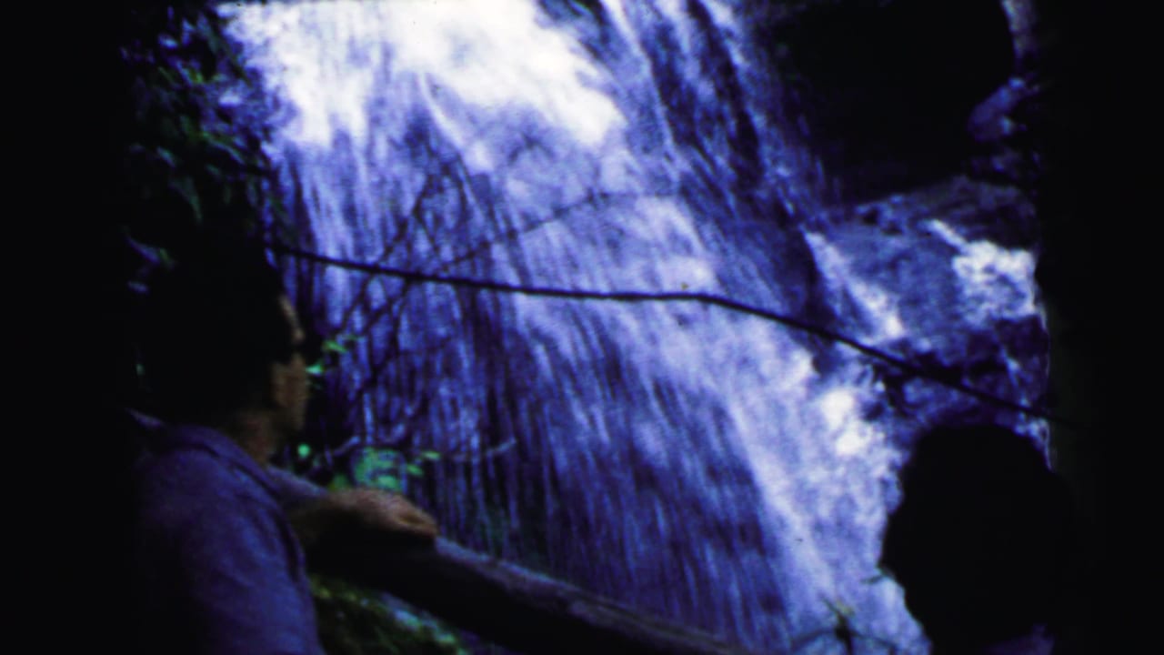 Two partially silhouetted figures standing beside a cascading waterfall, one in profile, green foliage on either side.