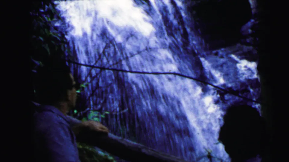 Two adults partially backlit against a bright cascading waterfall in 8mm home movie footage from an outdoor visit, circa 1964.