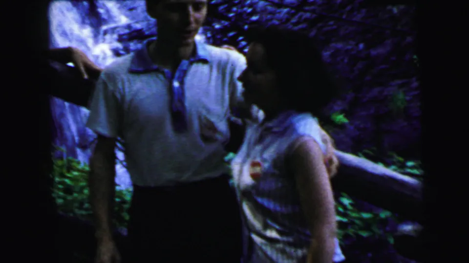Couple in 1960s casual wear conversing beside a waterfall, wooden railing in foreground; 8mm footage from Camden, New Jersey, 1964.