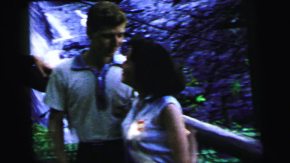 Close view of a couple in mid-1960s summer dress talking beside a waterfall; small round badge visible on woman's shirt; 8mm footage, circa 1964.
