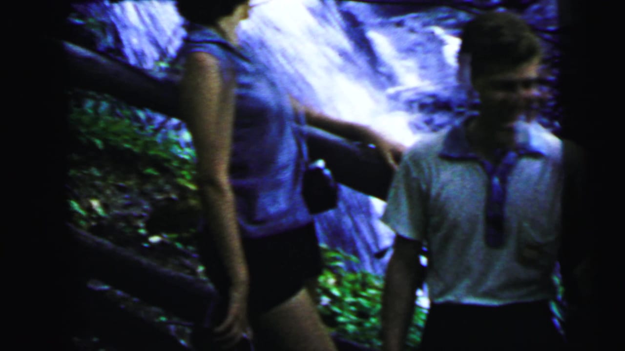 Young couple near a wooden railing with a cascading waterfall behind them, circa 1964 8mm home movie, strong blue-purple dye shift.