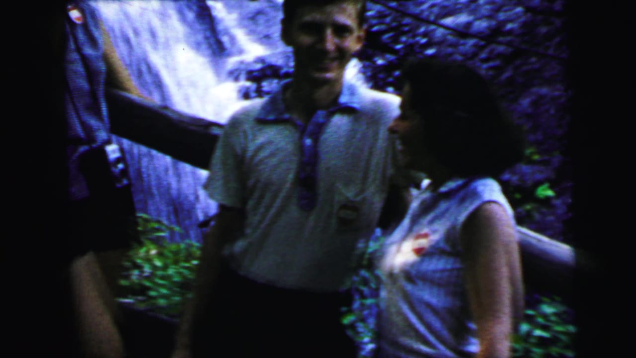 Young man in light polo shirt smiling toward camera, woman in sleeveless top at right, cascading waterfall and rocky cliff in background, circa 1964.