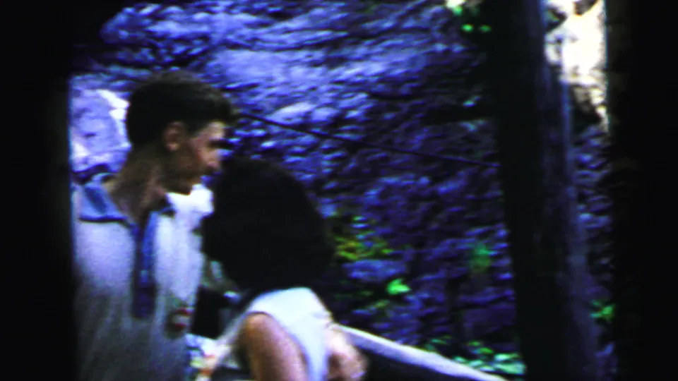 Close shot of young man looking downward, woman in white sleeveless top at right, waterfall cascading over dark rocks behind them, 8mm home movie circa 1964.