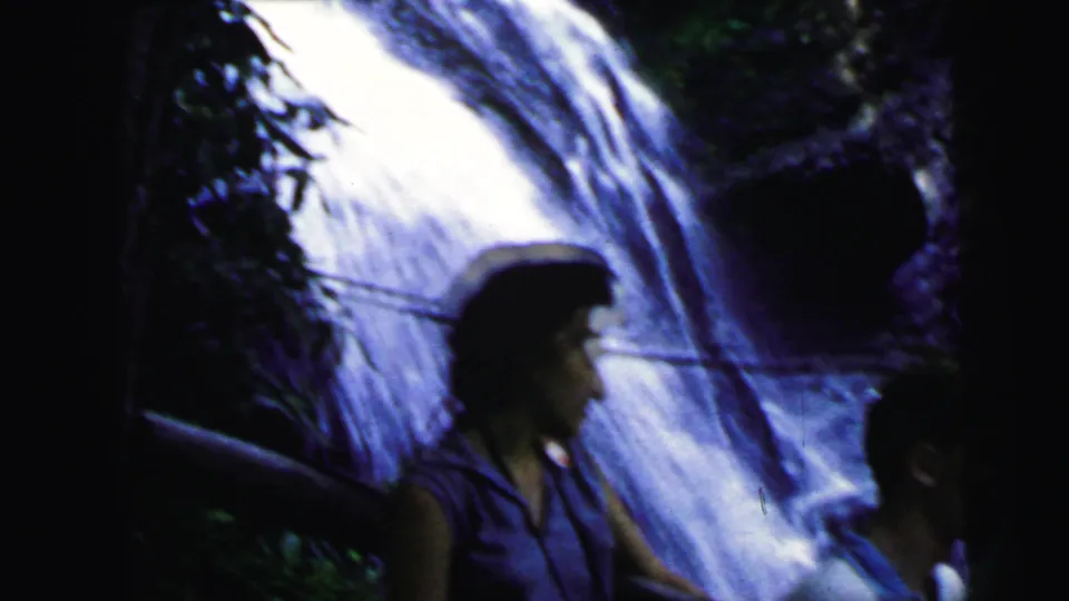 Woman with dark hair in white sleeveless top, man in polo shirt at left edge, waterfall cascading down a dark rocky face, 8mm home movie circa 1964.