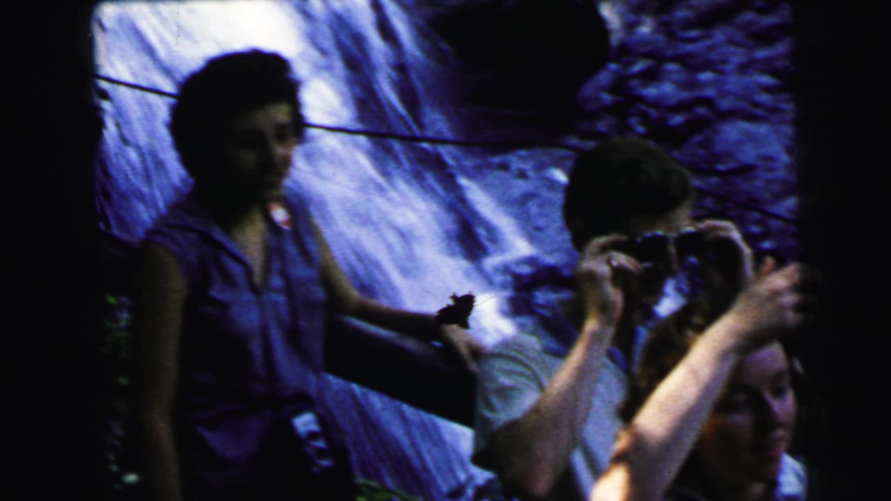 Three adults stand in front of a large rocky waterfall; one holds an object to their face.